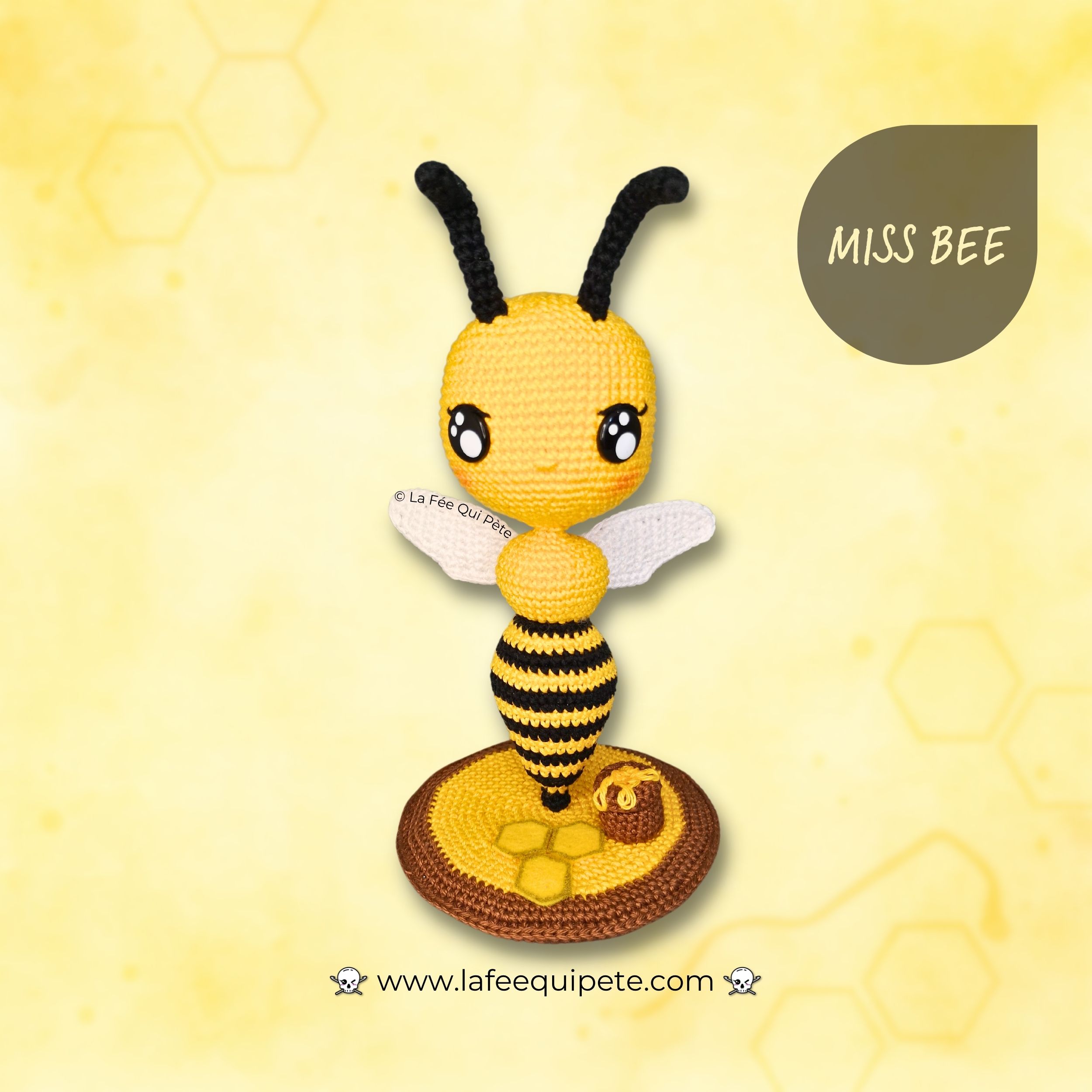 Abeille amigurumi complète posée sur son socle rond décoré, ailes blanches, antennes noires et visage expressif; fond jaune pastel à motifs d’alvéoles.